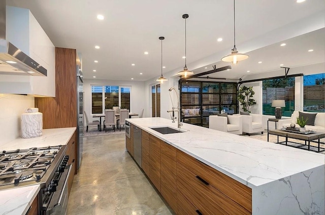 kitchen featuring wall chimney range hood, sink, appliances with stainless steel finishes, decorative light fixtures, and a large island with sink