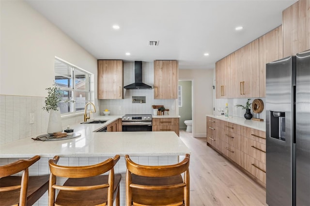 kitchen featuring sink, kitchen peninsula, stainless steel appliances, light hardwood / wood-style floors, and wall chimney range hood