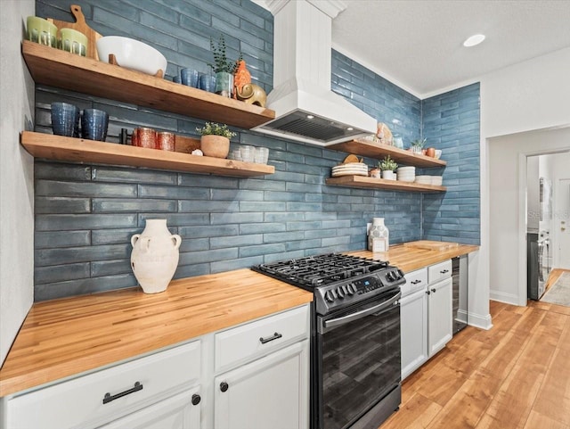 kitchen with butcher block counters, stainless steel gas range oven, custom range hood, white cabinets, and decorative backsplash