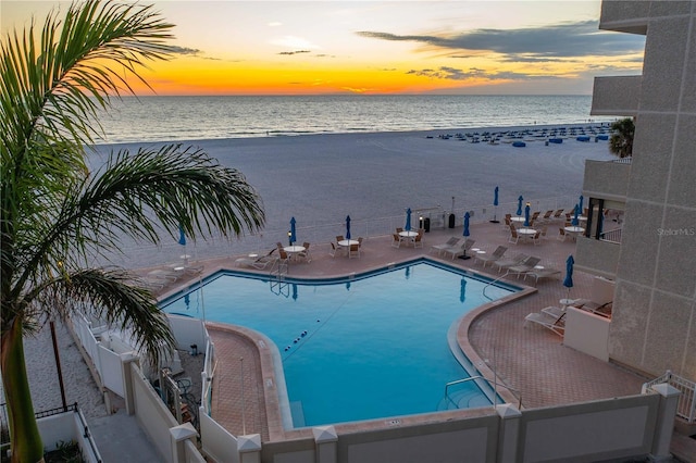 pool at dusk featuring a water view, a beach view, and a patio