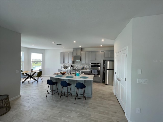 kitchen with gray cabinets, appliances with stainless steel finishes, tasteful backsplash, a breakfast bar area, and a center island with sink