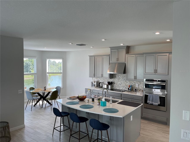 kitchen featuring sink, gray cabinets, double oven, backsplash, and an island with sink