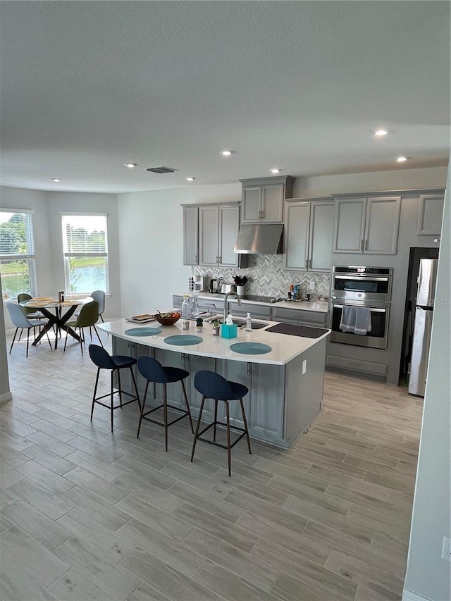 kitchen featuring appliances with stainless steel finishes, an island with sink, gray cabinetry, a kitchen breakfast bar, and decorative backsplash