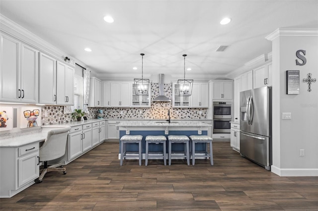 kitchen featuring appliances with stainless steel finishes, white cabinetry, a kitchen bar, a center island with sink, and wall chimney exhaust hood