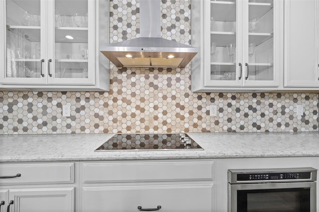 kitchen featuring wall chimney range hood, light stone counters, black electric stovetop, white cabinets, and oven