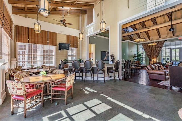 dining space featuring beamed ceiling, a towering ceiling, ceiling fan, and wood walls