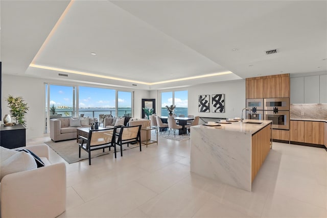 kitchen featuring sink, a water view, a tray ceiling, light stone countertops, and a center island with sink