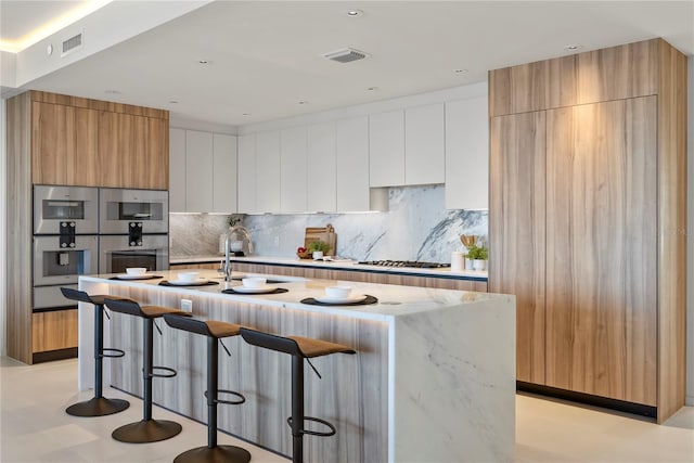 kitchen featuring tasteful backsplash, an island with sink, white cabinets, a kitchen bar, and light stone countertops