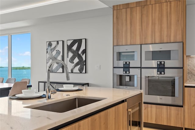 kitchen featuring sink, beverage cooler, light stone counters, a water view, and stainless steel double oven