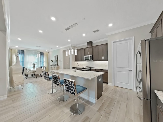 kitchen featuring a breakfast bar, appliances with stainless steel finishes, a kitchen island with sink, dark brown cabinets, and light stone counters