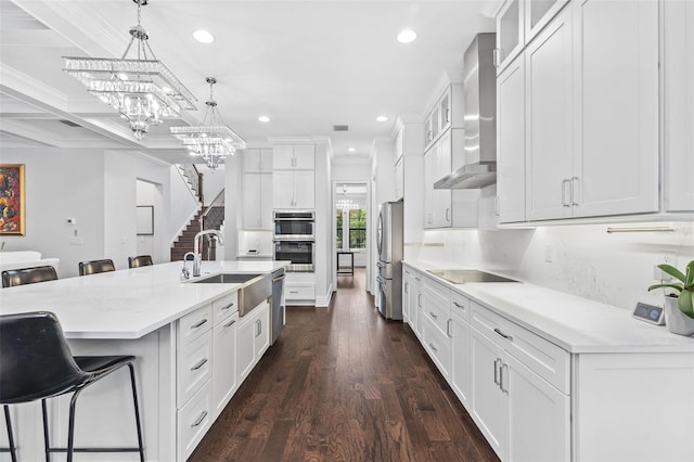 kitchen featuring a kitchen island with sink, stainless steel appliances, white cabinets, decorative light fixtures, and wall chimney exhaust hood