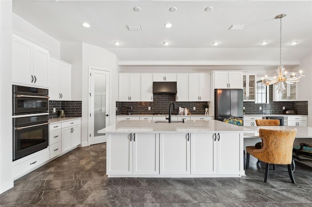 kitchen featuring stainless steel refrigerator, double oven, white cabinetry, sink, and a center island with sink