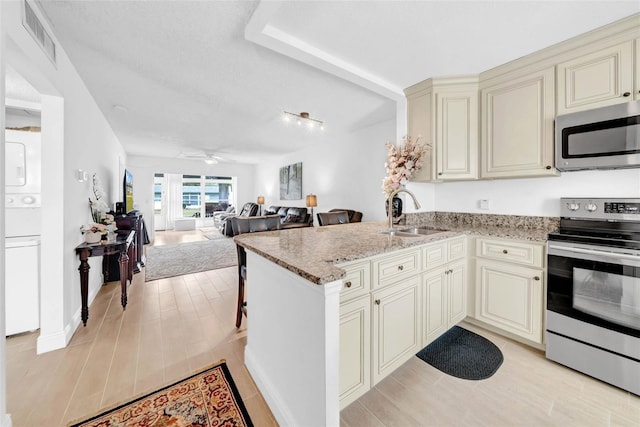 kitchen with appliances with stainless steel finishes, sink, stacked washer and clothes dryer, light stone counters, and cream cabinetry