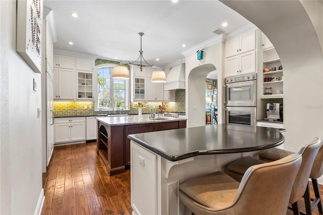 kitchen featuring premium range hood, double oven, an island with sink, white cabinets, and decorative light fixtures