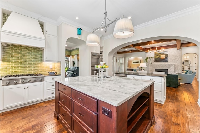 kitchen featuring light stone counters, custom range hood, an island with sink, decorative light fixtures, and stainless steel gas stovetop