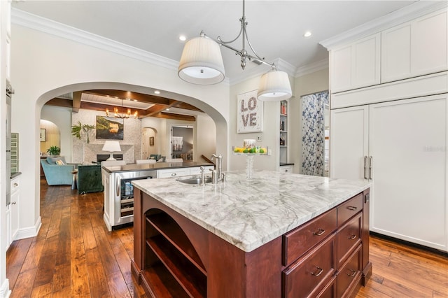 kitchen with arched walkways, open shelves, a kitchen island with sink, a sink, and beverage cooler