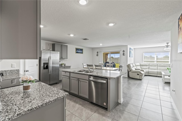 kitchen with a center island with sink, appliances with stainless steel finishes, light stone countertops, gray cabinetry, and a sink