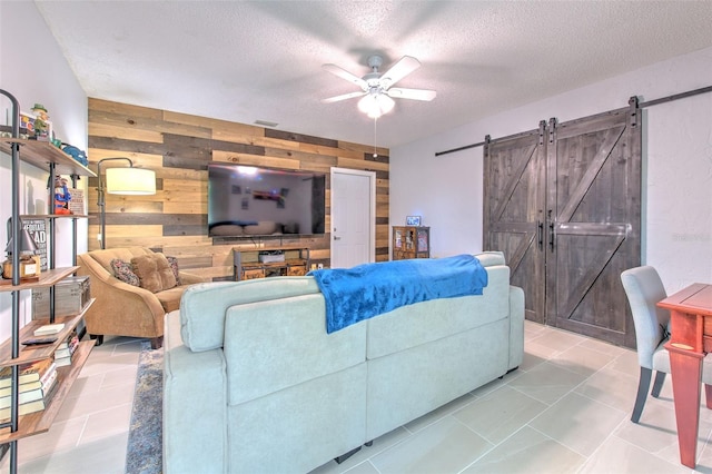 tiled living room featuring a barn door, wooden walls, a ceiling fan, an accent wall, and a textured ceiling