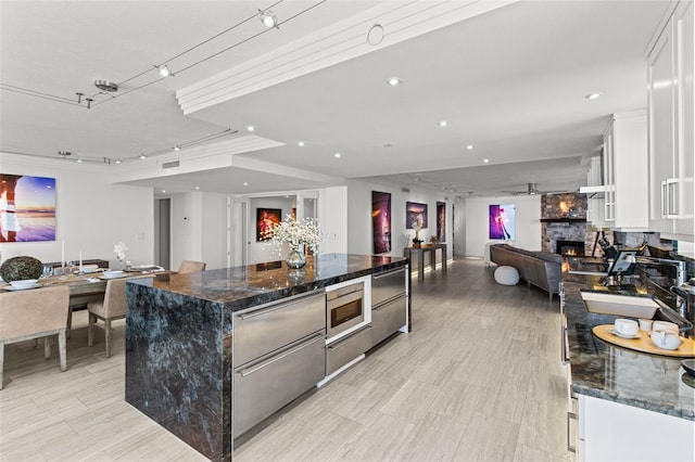 kitchen with a spacious island, stainless steel microwave, white cabinets, a stone fireplace, and dark stone counters