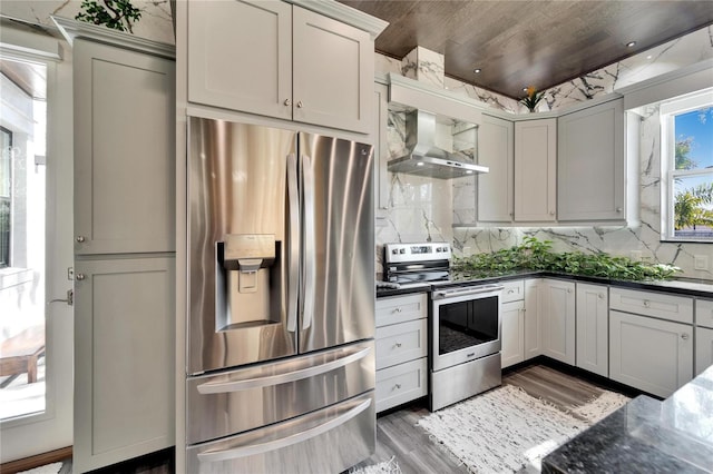 kitchen featuring wood-type flooring, decorative backsplash, dark stone counters, stainless steel appliances, and wall chimney exhaust hood