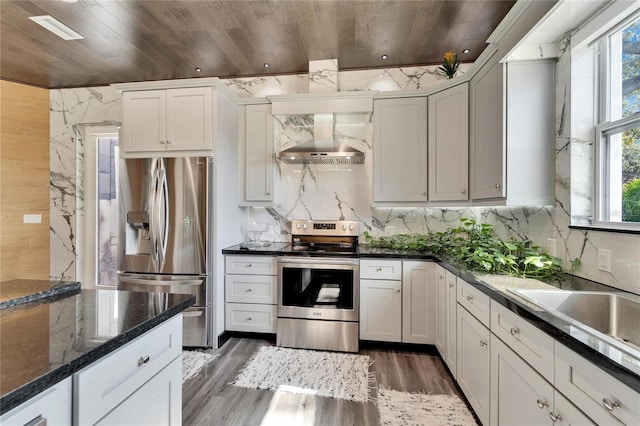 kitchen featuring wall chimney exhaust hood, wooden ceiling, dark stone countertops, stainless steel appliances, and white cabinets