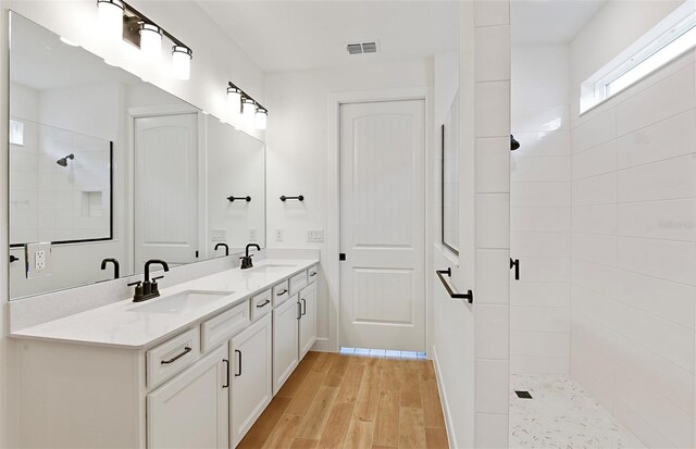 full bath with a sink, a tile shower, double vanity, and wood finished floors