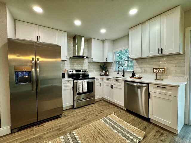 kitchen with sink, wall chimney range hood, light wood-type flooring, stainless steel appliances, and white cabinets