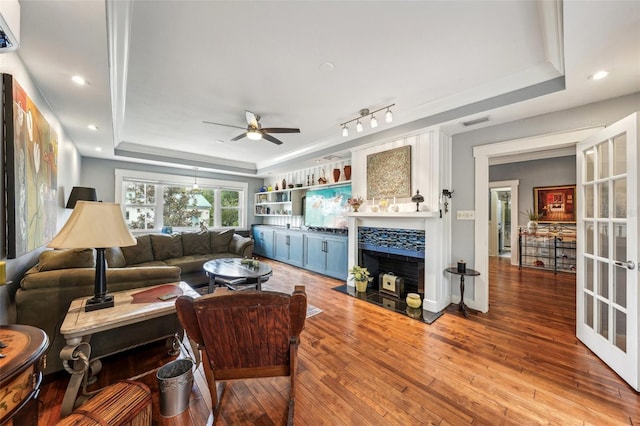 living room featuring a stone fireplace, a raised ceiling, ceiling fan, and light wood-type flooring