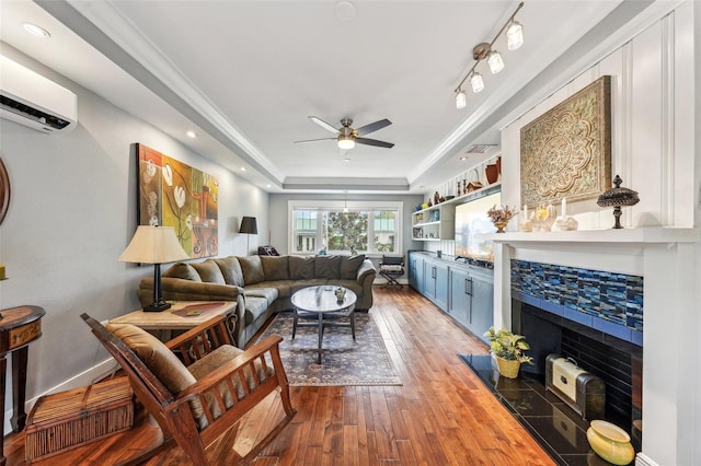 living room featuring a wall mounted air conditioner, wood-type flooring, track lighting, a raised ceiling, and ceiling fan