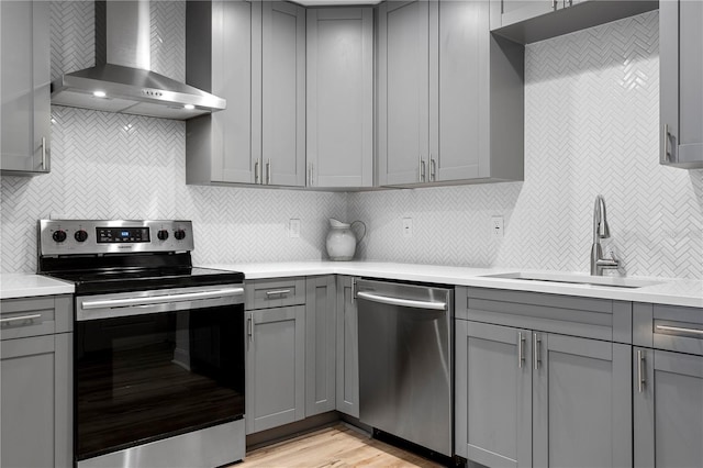 kitchen with gray cabinetry, a sink, light countertops, wall chimney range hood, and appliances with stainless steel finishes