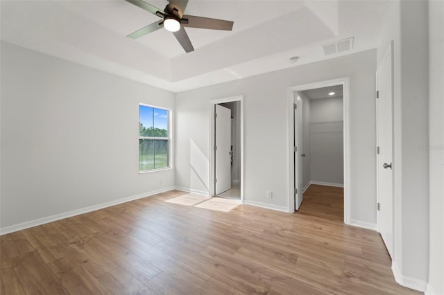 unfurnished bedroom with a spacious closet, light hardwood / wood-style flooring, a tray ceiling, a closet, and ceiling fan