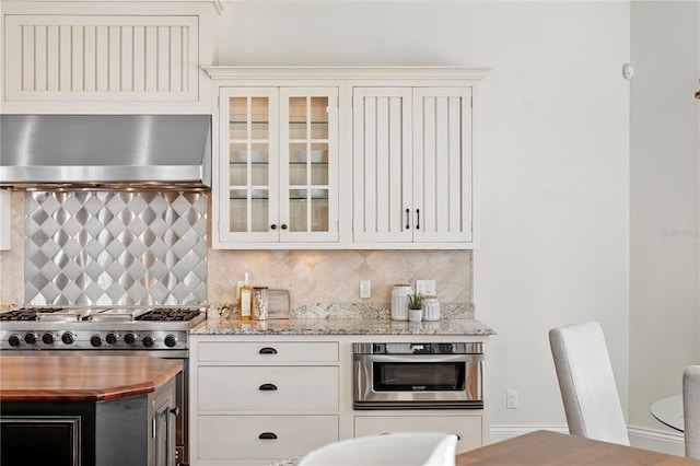 kitchen featuring butcher block countertops, decorative backsplash, glass insert cabinets, appliances with stainless steel finishes, and wall chimney range hood