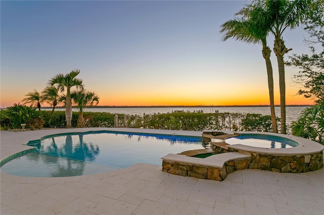 pool at dusk with a patio area, an outdoor pool, an in ground hot tub, and a water view