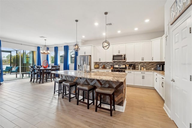 kitchen featuring white cabinetry, hanging light fixtures, stainless steel appliances, light stone countertops, and a center island with sink