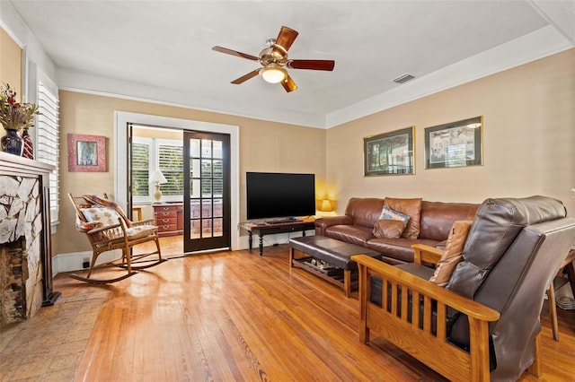 living room with crown molding, a stone fireplace, ceiling fan, and light wood-type flooring