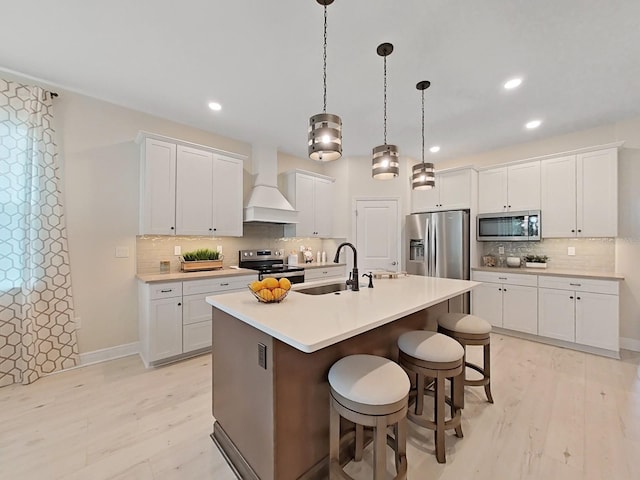 kitchen featuring decorative light fixtures, appliances with stainless steel finishes, an island with sink, custom range hood, and white cabinets