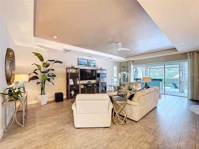 living room with baseboards, a raised ceiling, a ceiling fan, a textured ceiling, and light wood-type flooring