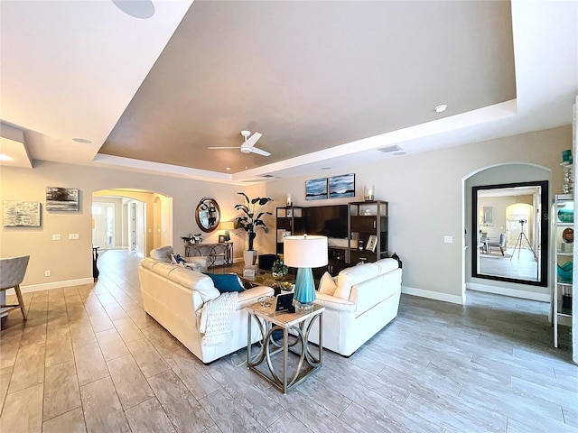 living area featuring arched walkways, a raised ceiling, a ceiling fan, and light wood-style floors