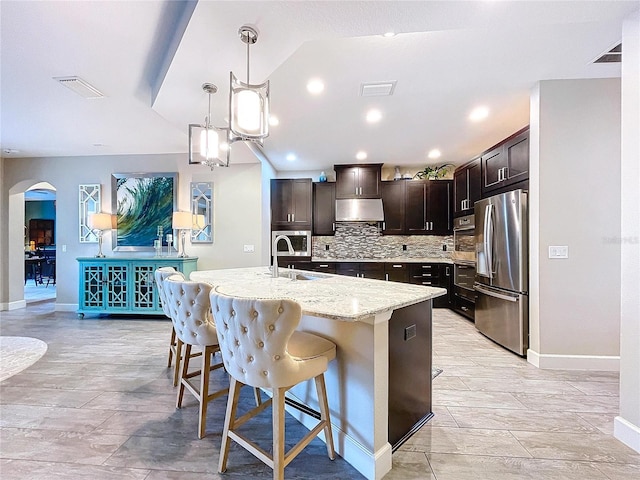 kitchen featuring arched walkways, decorative backsplash, freestanding refrigerator, a sink, and under cabinet range hood