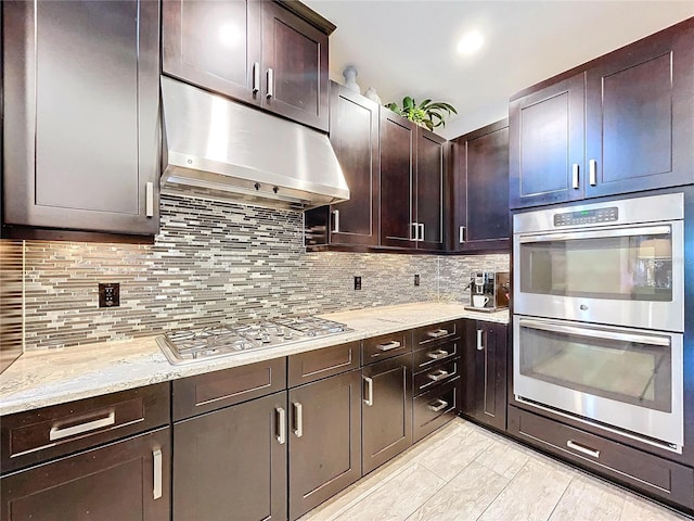 kitchen featuring light stone counters, stainless steel appliances, backsplash, dark brown cabinets, and under cabinet range hood