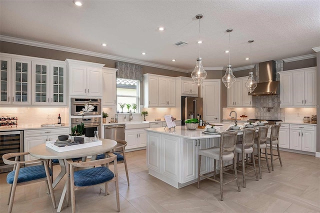 kitchen featuring pendant lighting, wall chimney range hood, stainless steel appliances, a center island with sink, and beverage cooler