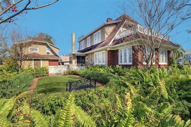 view of front facade with a balcony, brick siding, fence, a front lawn, and a chimney