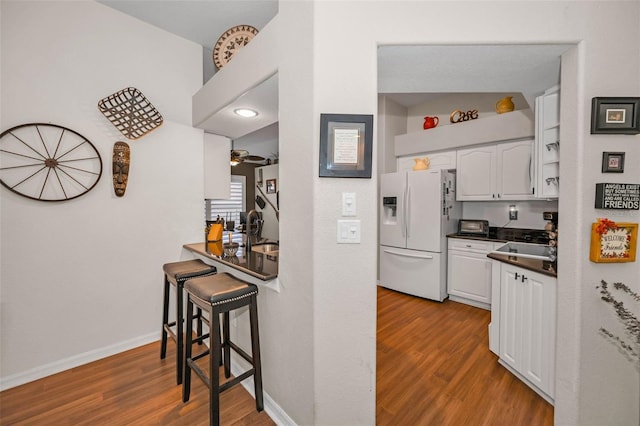 kitchen with white refrigerator with ice dispenser, dark countertops, white cabinets, a sink, and wood finished floors