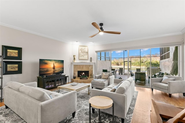 living room with a tiled fireplace, crown molding, ceiling fan, and light wood-type flooring