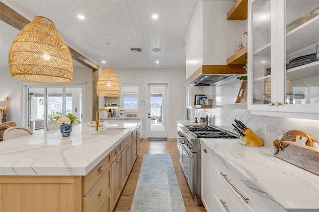 kitchen with high end range, range hood, light stone counters, an island with sink, and white cabinets