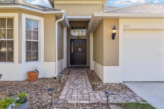 view of exterior entry with a garage, roof with shingles, and stucco siding