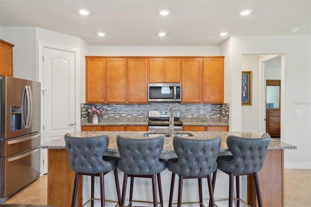7580 Pool Compass Loop, Wesley Chapel, FL 33545 - kitchen with brown cabinetry, light tile patterned flooring, a center island with sink, backsplash, and stainless steel appliances