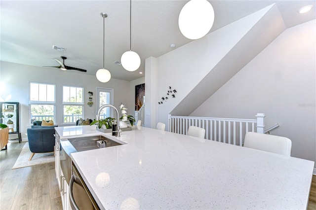 kitchen with dishwasher, hanging light fixtures, light stone counters, light hardwood / wood-style floors, and a center island with sink