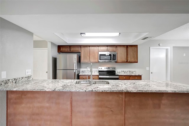 kitchen with sink, stainless steel appliances, light stone counters, a tray ceiling, and kitchen peninsula