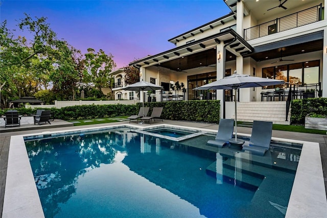 pool at dusk featuring a patio area, ceiling fan, fence, and a pool with connected hot tub